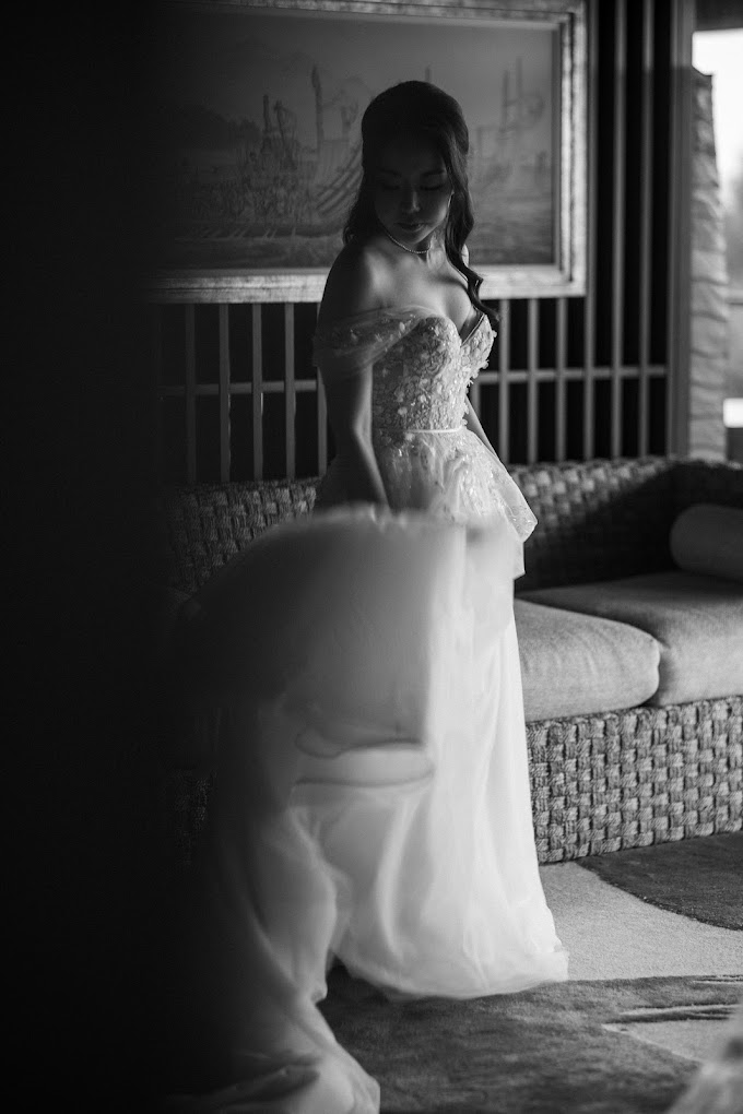 Woman in a strapless dress stands indoors near a couch, looking down while adjusting her gown. Black and white image.