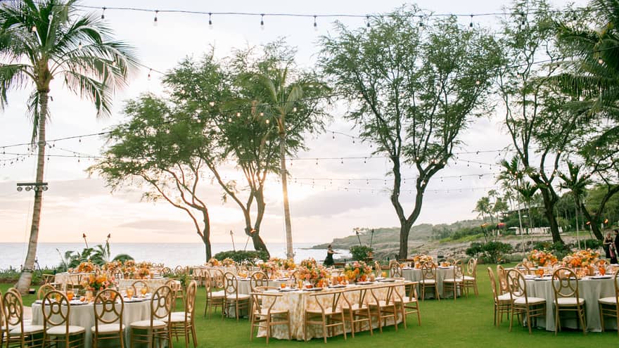 Outdoor wedding reception setup on a grassy area by the ocean, featuring decorated round tables, wooden chairs, string lights, and palm trees under a clear sky.