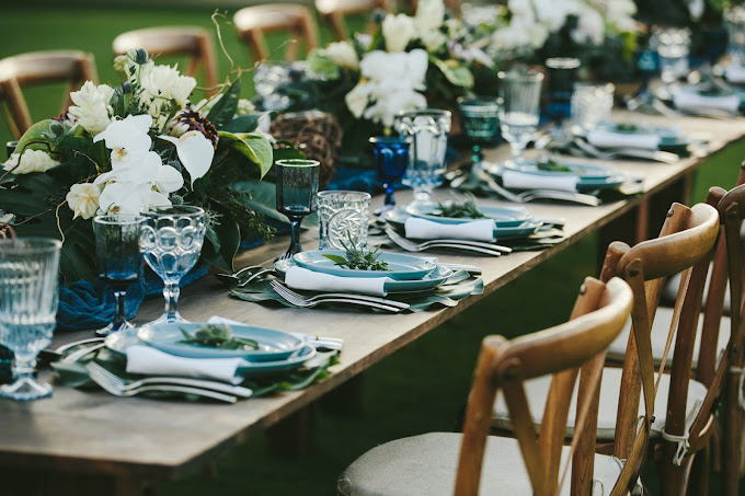 A long dining table is set with elegant dinnerware, glassware, and floral centerpieces. Wooden chairs line both sides of the table.