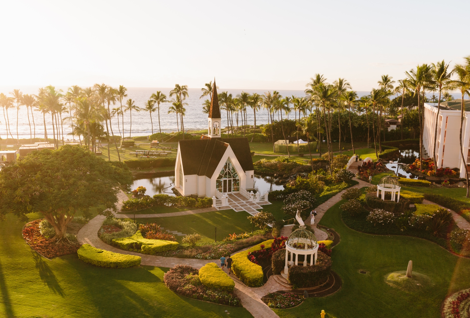 A picturesque chapel surrounded by lush gardens and palm trees near the ocean at sunset, with pathways leading through the landscaped grounds.