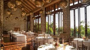 Spacious dining area with stone walls, large windows, wooden ceiling beams, and tables set for a meal. Spherical pendant lights hang from the ceiling.