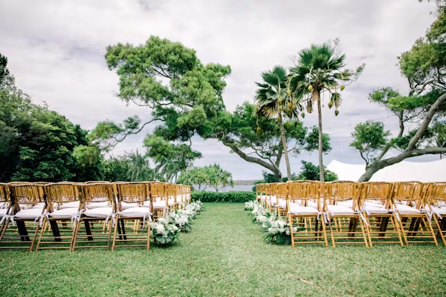 Outdoor wedding setup with rows of wooden chairs, greenery, and white flowers on a grassy lawn, with trees in the background under a cloudy sky.