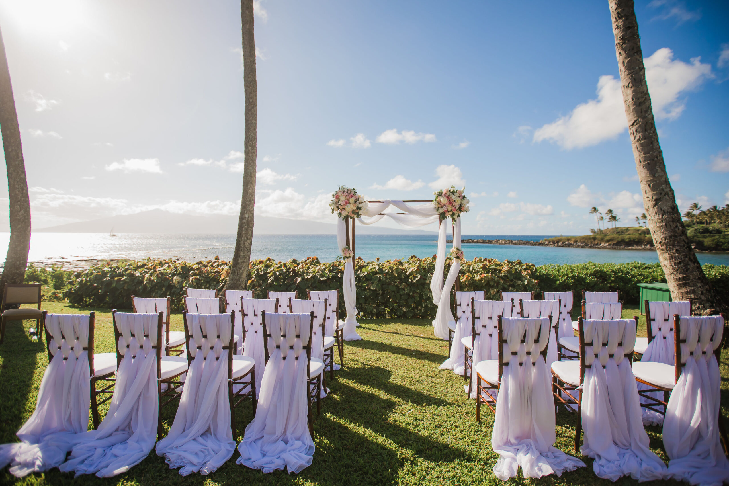 Outdoor wedding setup with white draped chairs facing an arch adorned with flowers, positioned between two palm trees overlooking the ocean under a clear blue sky.