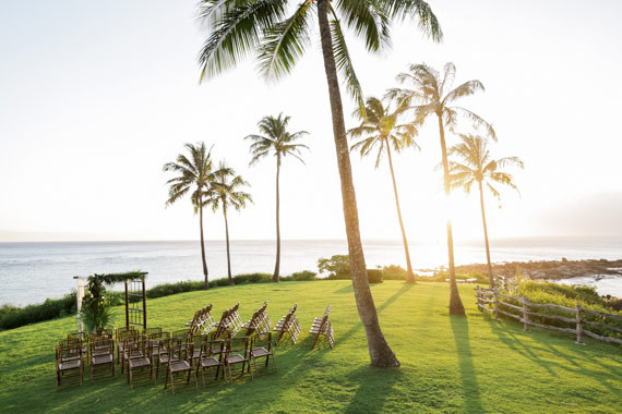 Outdoor wedding setup on a grassy area by the sea with rows of wooden chairs and a simple arch, framed by palm trees under a clear sky.