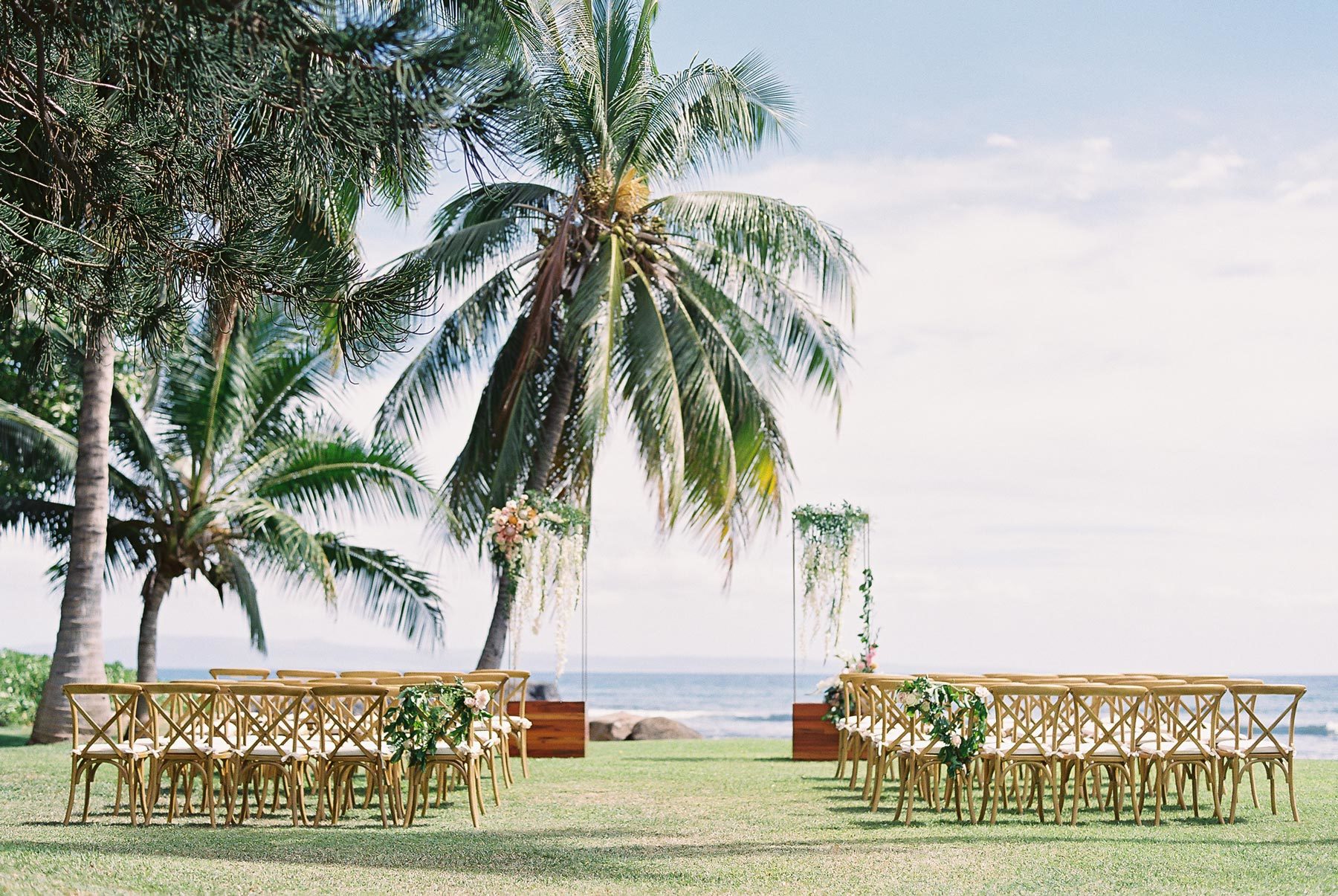 Outdoor wedding setup on grass with wooden chairs facing the ocean, framed by palm trees and floral arrangements.